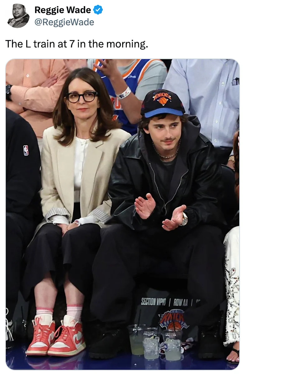 Two people sitting courtside, one in a leather jacket and cap, clapping. The other in glasses and casual attire, with red sneakers