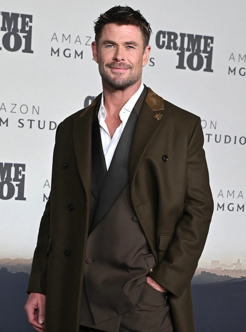 Man in a stylish suit and overcoat poses at a Crime 101 event backdrop