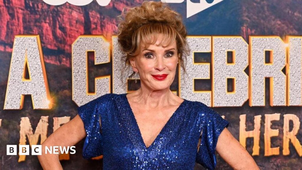 Beverley Callard in a blue dress with her hands on her hips, standing in front of a sign for I'm a Celebrity... South Africa