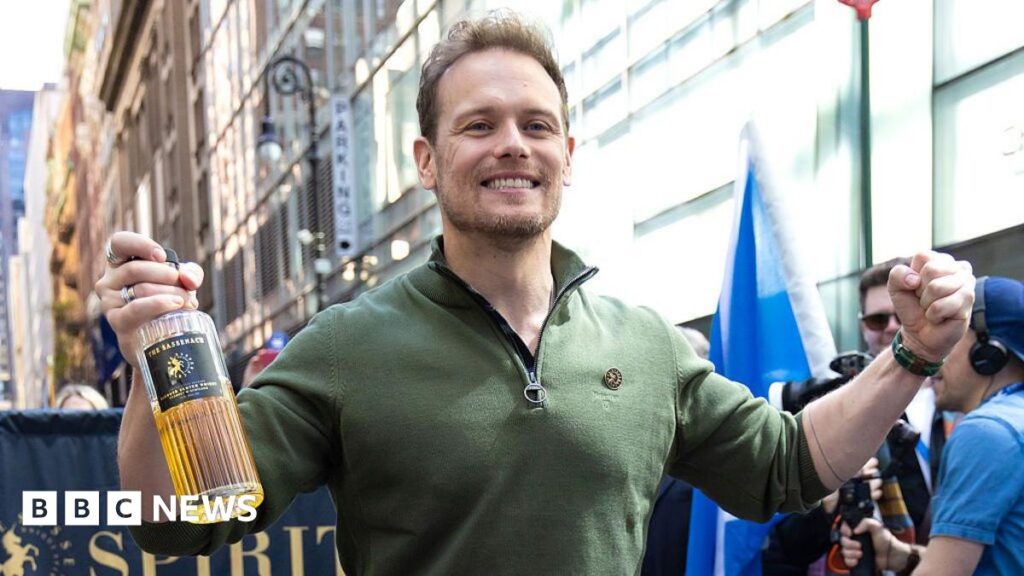 Outlander star Sam Heughan leads New York Tartan Day parade A man in a green top, smiling and holding a bottle of whisky in his hand, with a column of marchers behind him
