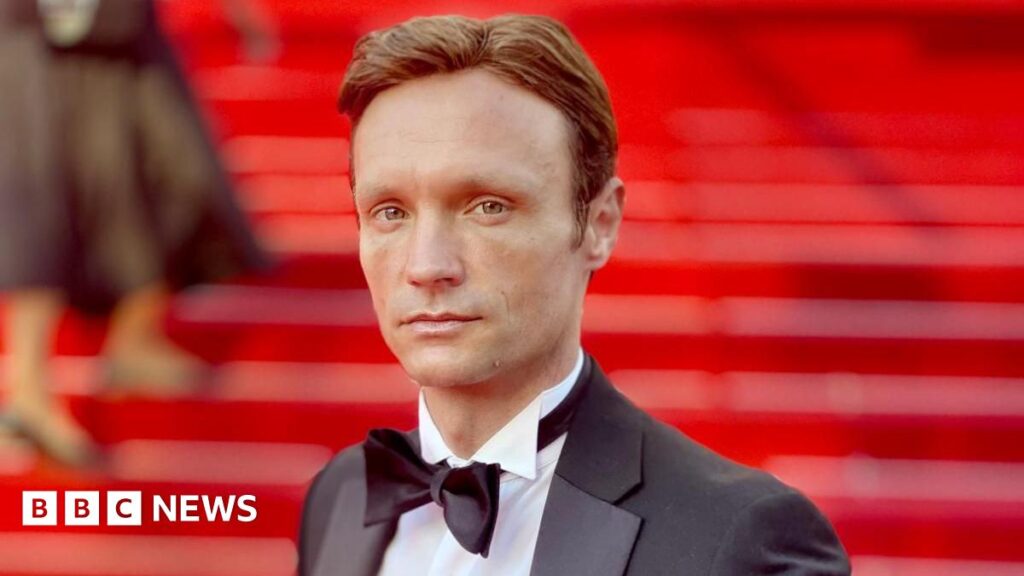 Hans Obma stood on a red carpet. Red stairs can be seen behind him, he wears a black suit jacket, white shirt and black bow tie. He has blue eyes and brown hair and looks side on down the lens.