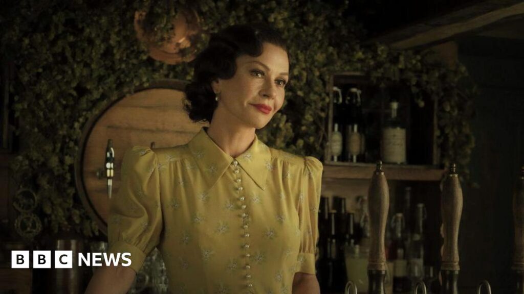 Catherine Zeta Jones in a yellow dress. She has short dark hair and is standing behind an old style bar.