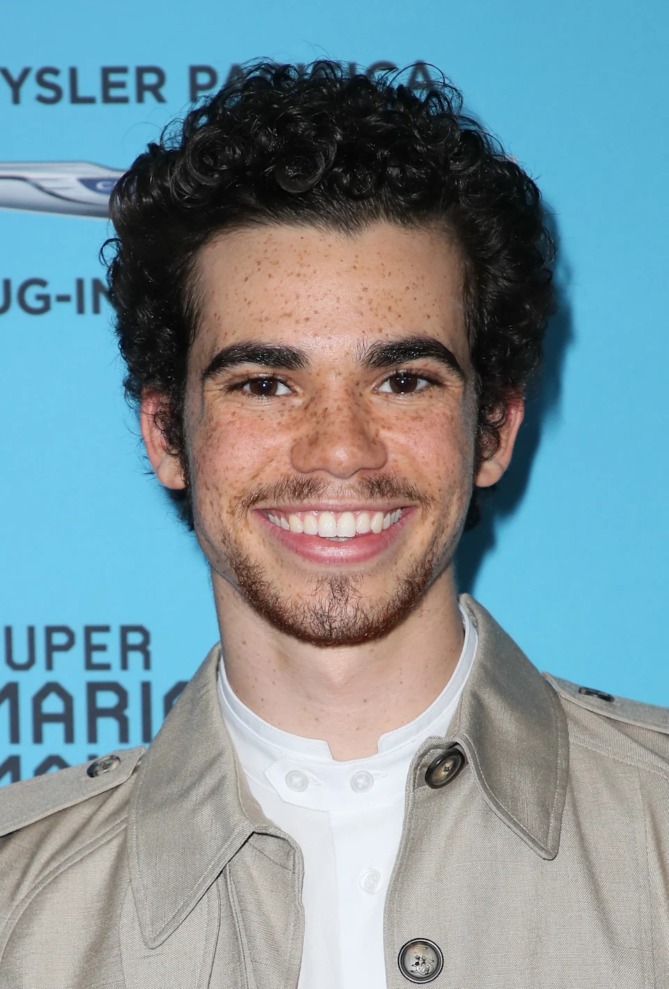 A smiling person with curly hair wears a stylish trench coat over a white shirt at a public event