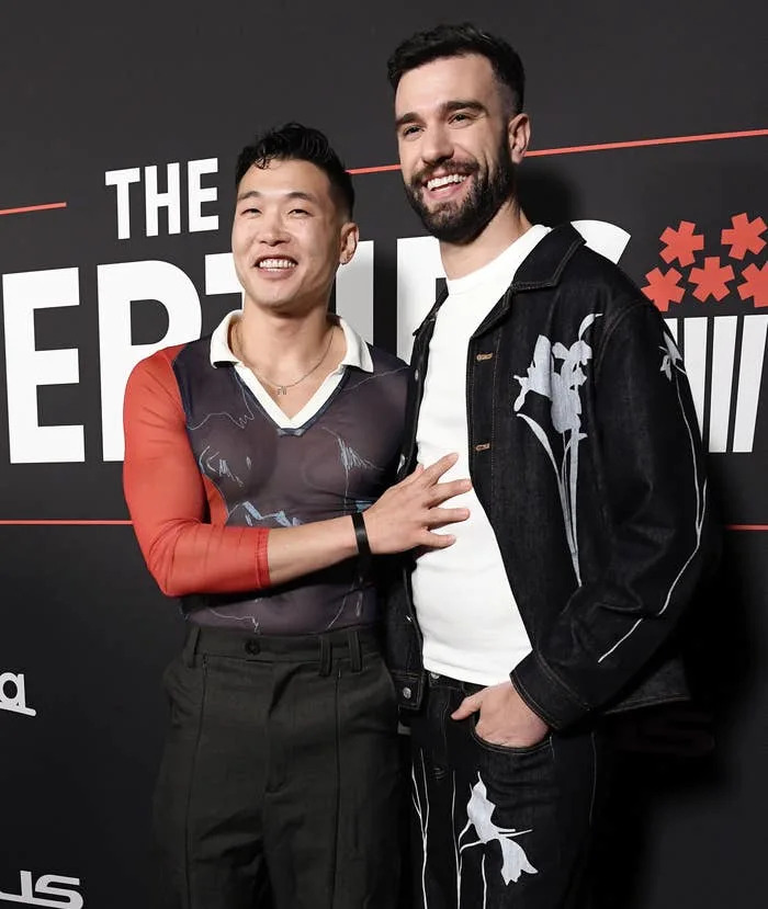 Two people pose together on a red carpet. One wears a stylish long-sleeve top with artistic designs, and the other wears a matching denim jacket and pants