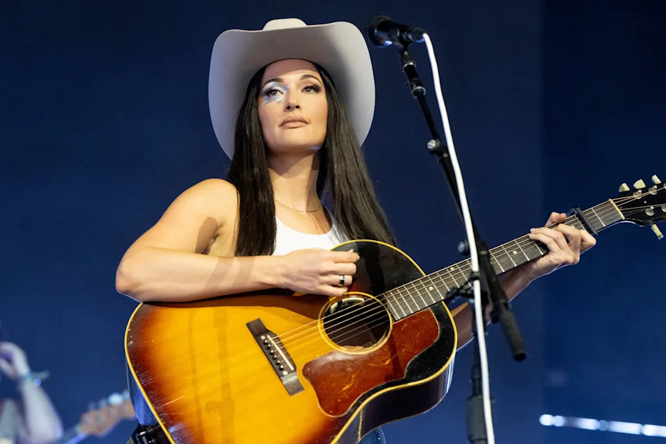 Kacey Musgraves wearing a cowboy hat and a tank top performs on stage with an acoustic guitar