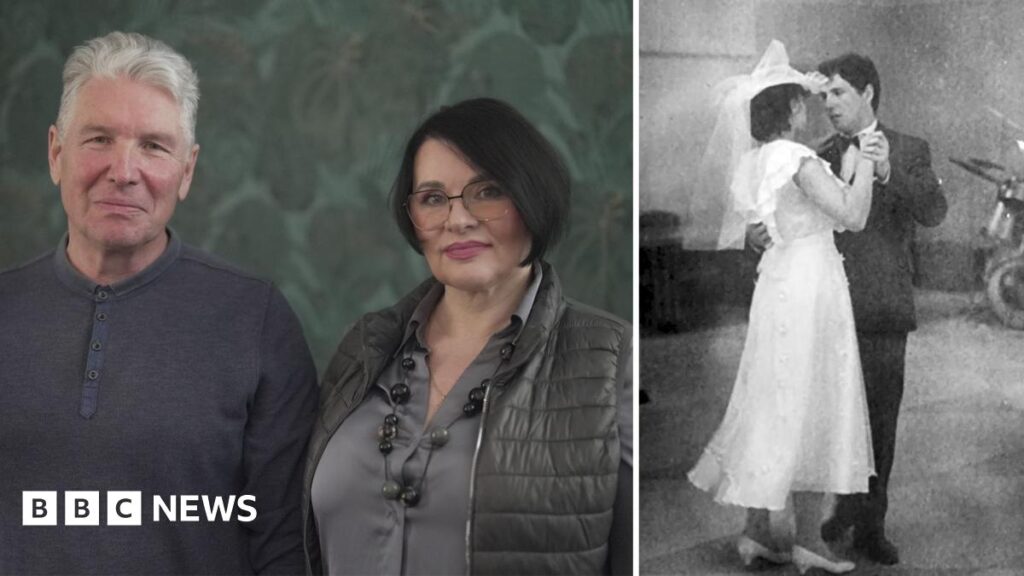 Iryna and Serhiy photographed in 2026, looking at the camera and smiling. Iryna has chin-length dark hair and glasses and is holding a pink flower. Serhiy has grey hair and is holding up a photo of them side-by-side at their wedding.