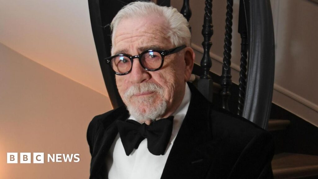 A dark wooded, stylish room is the backdrop for a dramatic moody shot of Alan Cumming, in a tartan blazer, brown tweed waistcoat and denim shirt, lifting a glass of whisky to his lips.