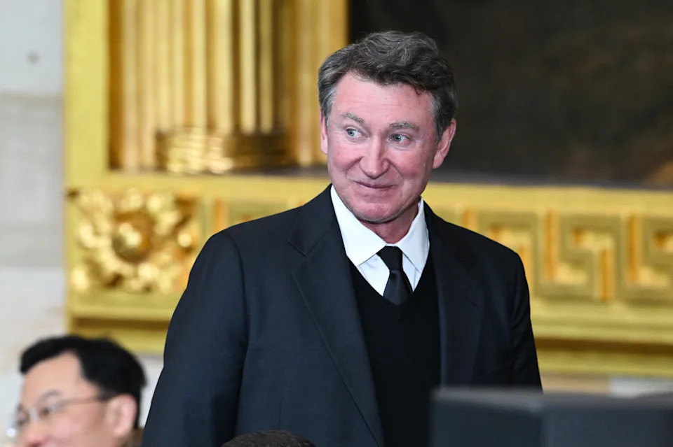 Wayne Gretzky in a formal suit and tie stands indoors, smiling, with ornate decor in the background