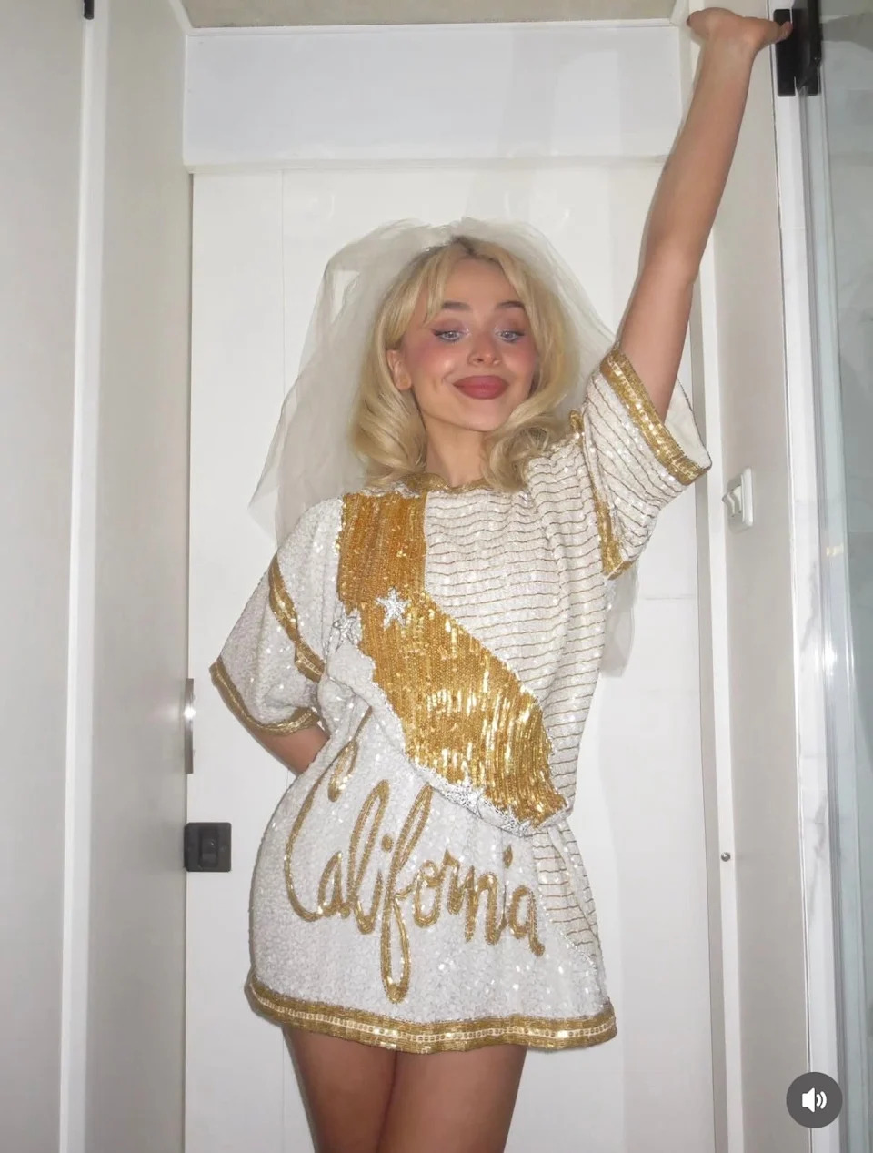 Sabrina Carpenter in a sequined mini dress with "California" text, wearing a veil, poses energetically with one arm raised