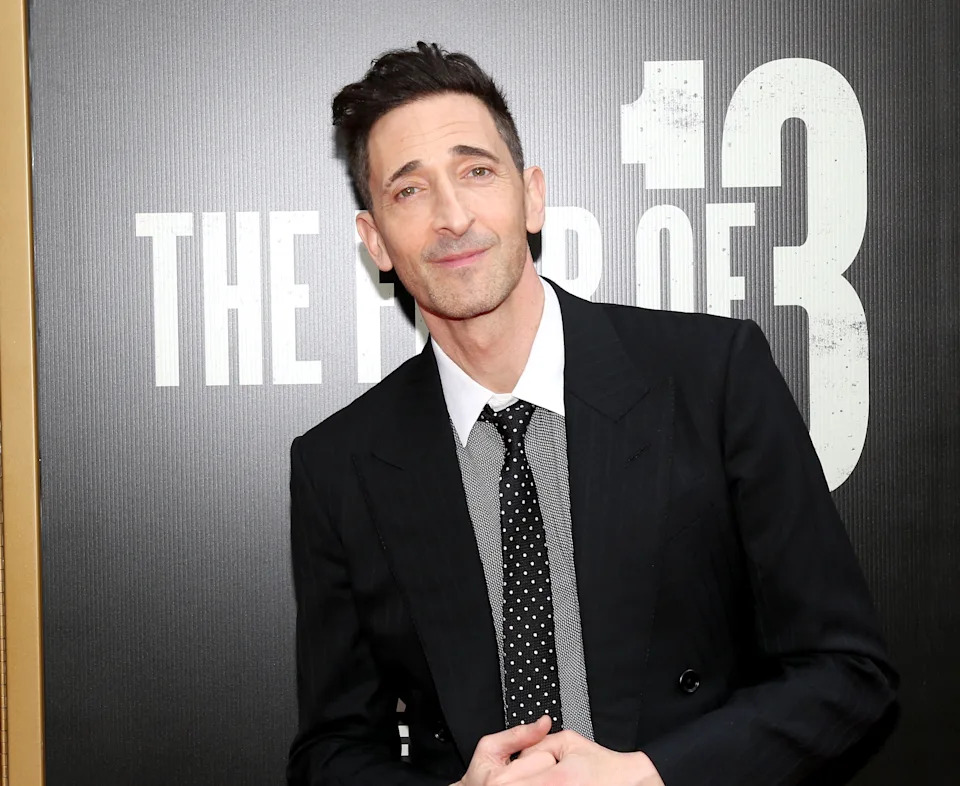 A person in a stylish suit and polka dot tie poses in front of a backdrop with bold text