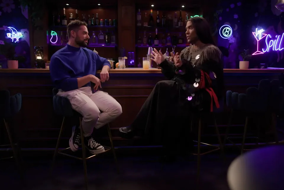 Two people having a conversation at a bar with neon signs. One wears a blue top and white pants; the other wears a black outfit with shiny accents
