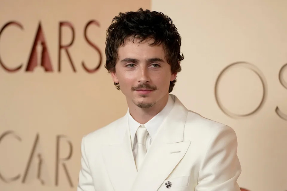 Person in a white suit with a pin on the lapel, attending a formal event with an Oscars backdrop