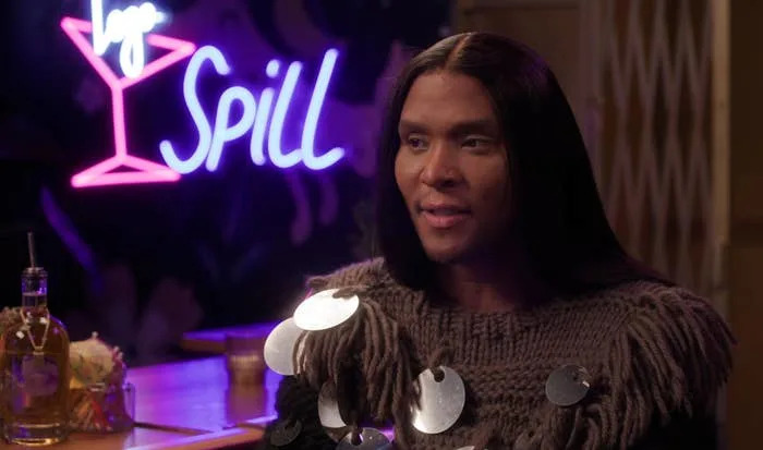 A person indoors wearing a textured sweater with large disc embellishments, sitting at a bar. Neon "Spill" sign in the background