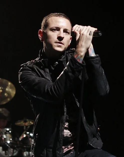 A person on stage passionately sings into a microphone, wearing a casual black jacket with visible tattoos