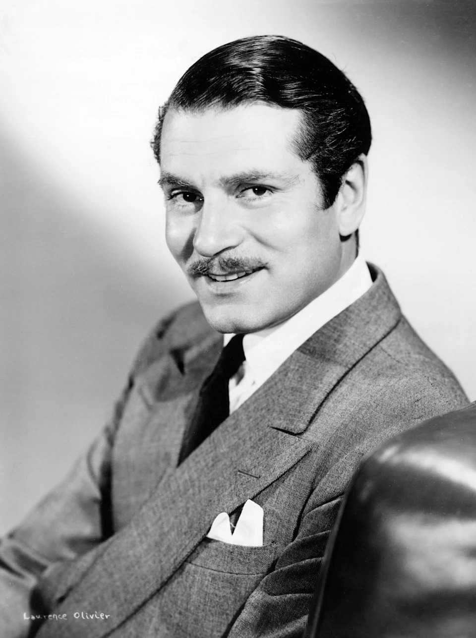 Laurence Olivier in a classic suit with slicked-back hair and a mustache, sitting and smiling directly at the camera