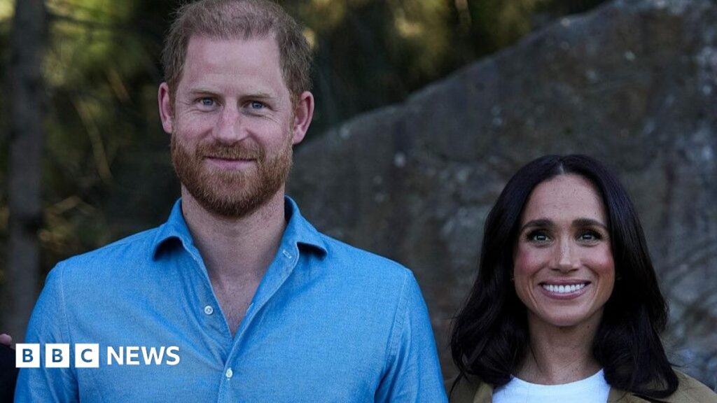 Harry stands next to Meghan. He is wearing a light blue shirt. They are both smiling.