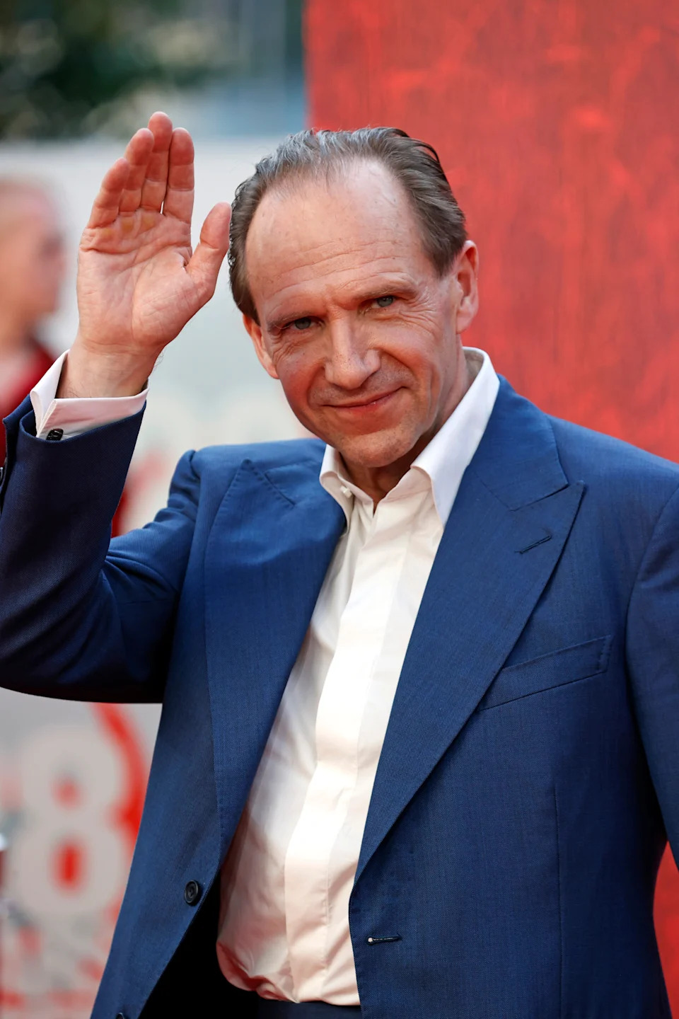 Person in a formal blue suit waves on the red carpet, smiling at the camera