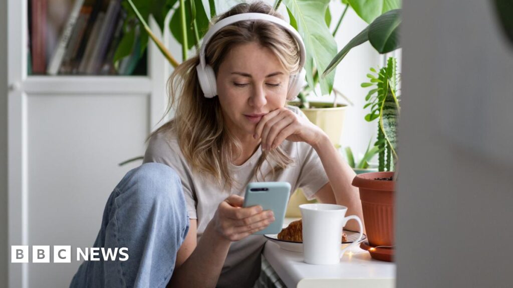 A stock shot of a young blonde woman wearing white headphone and looking at her phone.