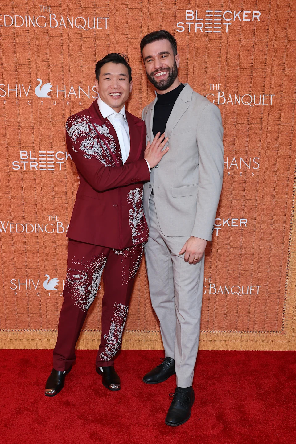 Two people smile on a red carpet. One wears a patterned suit, the other a plain suit. They stand in front of a backdrop with event sponsors