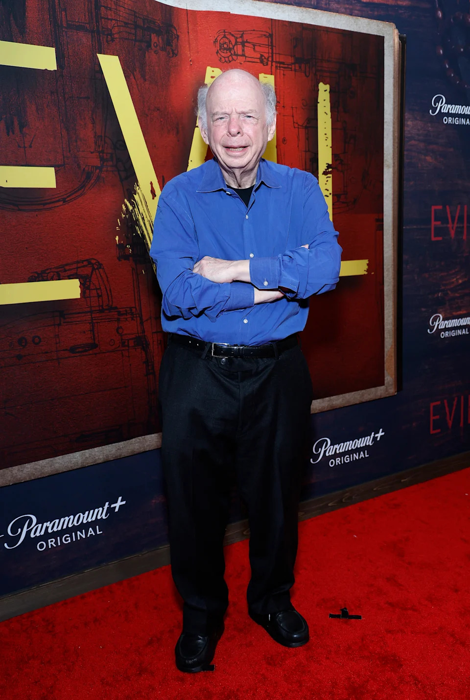 Person on red carpet in casual attire with a button-up shirt and black trousers, standing in front of an event backdrop