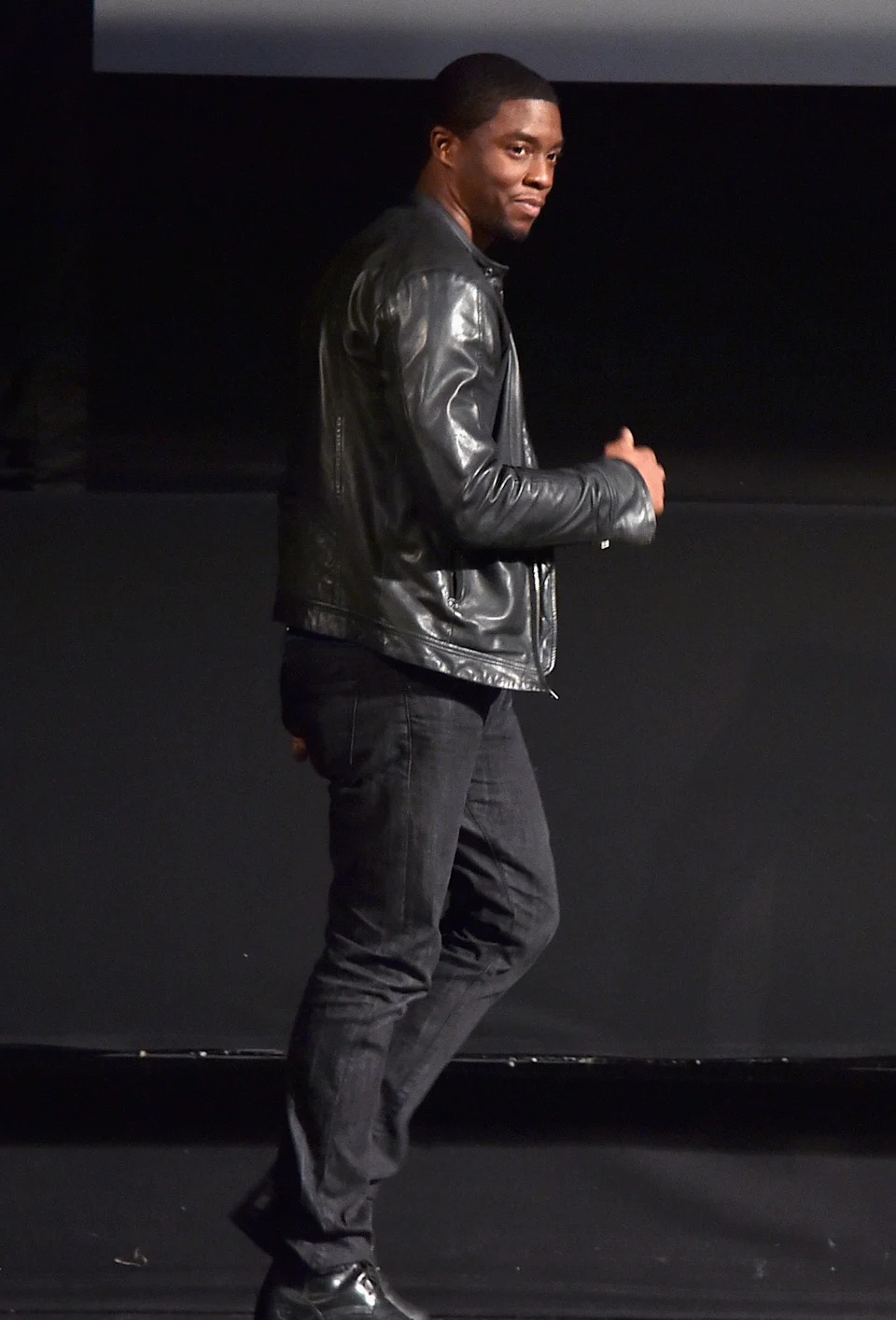 Chadwick Boseman in a stylish leather jacket and jeans is walking on stage, looking back with a confident expression