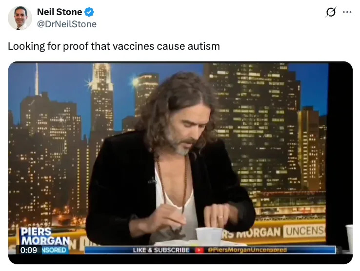 A man with long hair is seated at a table on a news show, appearing to search through papers, with a cityscape backdrop