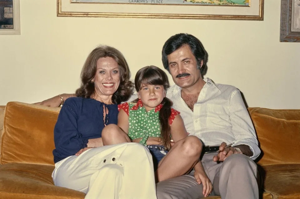 A family of three sits on a couch, with the child in the middle. The adults wear semi-casual 1970s attire