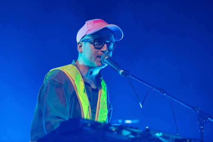 Alexis Taylor of Hot Chip performs on stage, wearing a pink cap, high vis jacket and shirt, while playing the keyboard and singing into a microphone