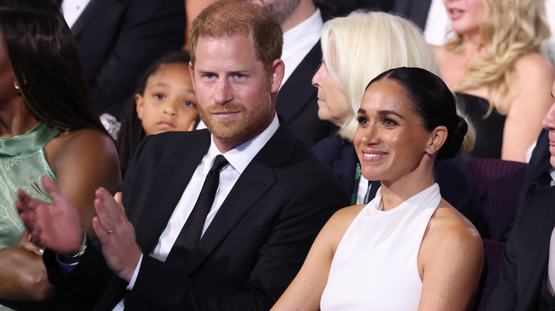 Prince Harry applauding in a suit next to a smiling Meghan Markle in a white dress