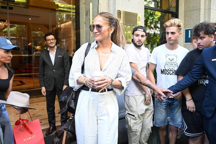 Jennifer Lopez, dressed in a striped blouse and white trousers, holds a coffee cup as she stands outside surrounded by fans and onlookers.