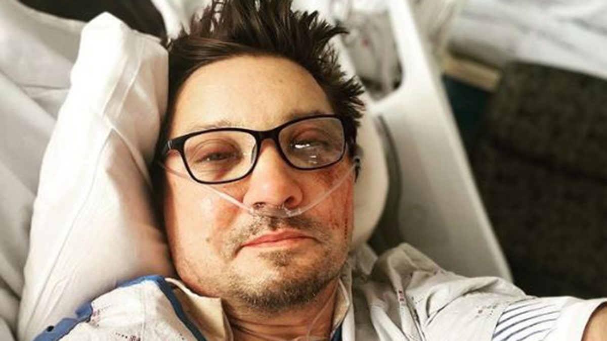 Jeremy Renner smiling in hospital bed wearing dark rimmed glasses and hospital gown with breathing tube in nose