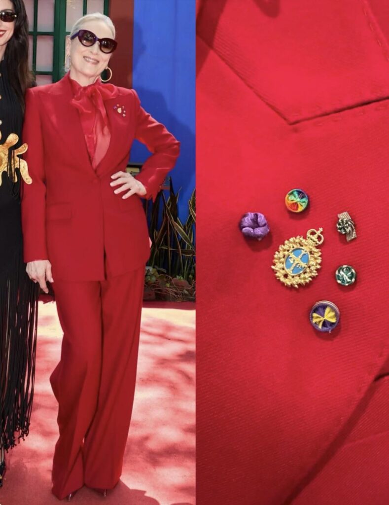Meryl Streep wearing her personal brooches including purple ribbons from her 2010 National Medal of the Arts, rainbow rosette from her 2011 Kennedy Center Honor, a gold cross on blue from the Princess of Asturias Awards, and green-and-white rosettes from the L'Ordre des Arts et des Lettres