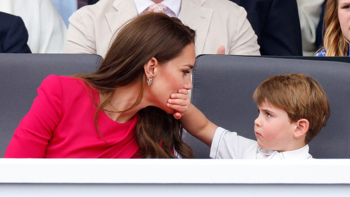 Prince Louis of Cambridge covering Catherine, Duchess of Cambridge's mouth with his hand at an outdoor event.