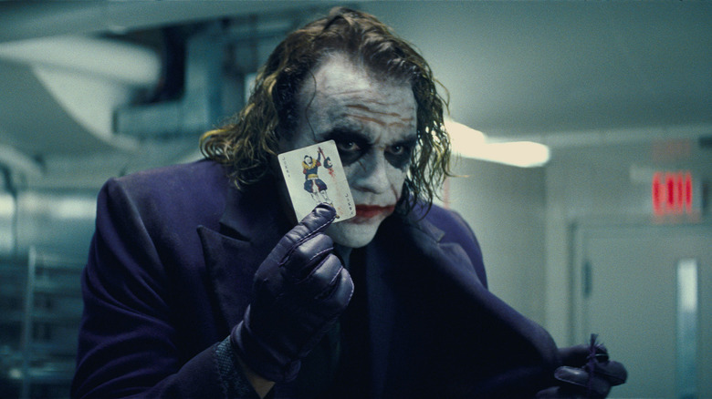 Heath Ledgers as the Joker holds up a card in The Dark Knight.