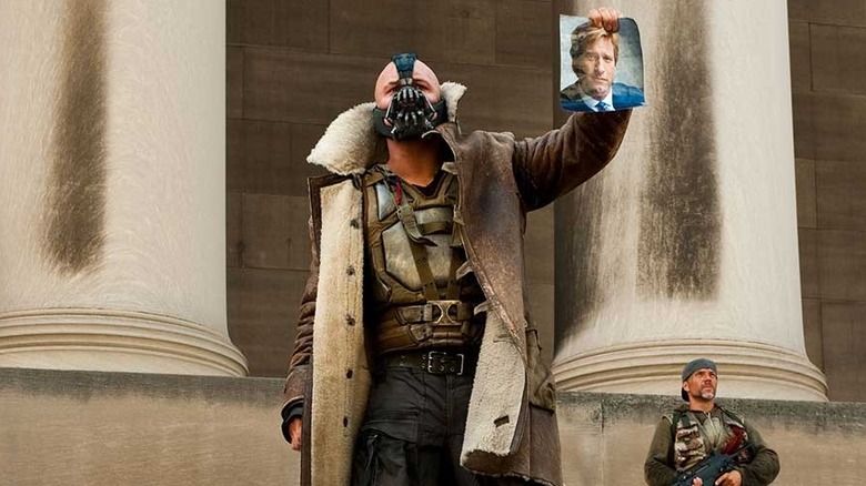 Tom Hardy as Bane, holding a photo of Aaron Eckhart's Harvey Dent in The Dark Knight Rises.