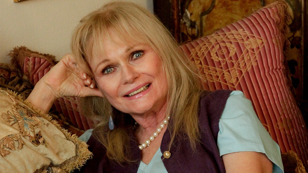 Valerie Perrine smiling seated on a couch in her Los Angeles home