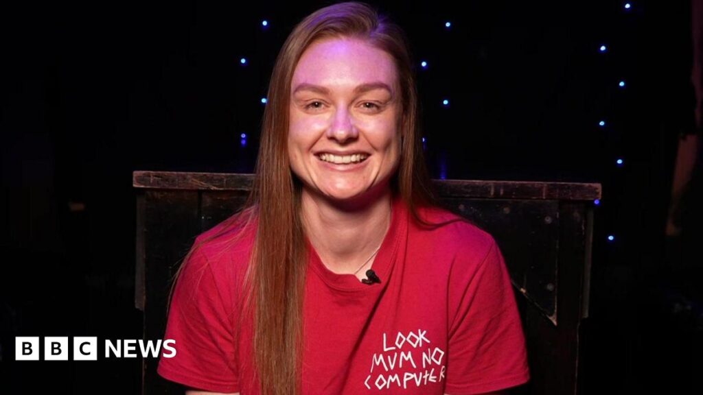 Sister of Peterborough-born Eurovision star bursting with pride Jodie Bartle is sitting in a dark room which has blue fairy lights hanging on the wall and a table. Bartle has long hair and she is wearing a red T-shirt.