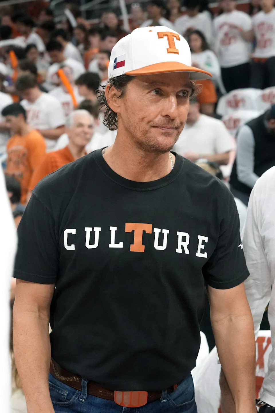 Man in "Culture" T-shirt and cap at a sports event surrounded by other attendees