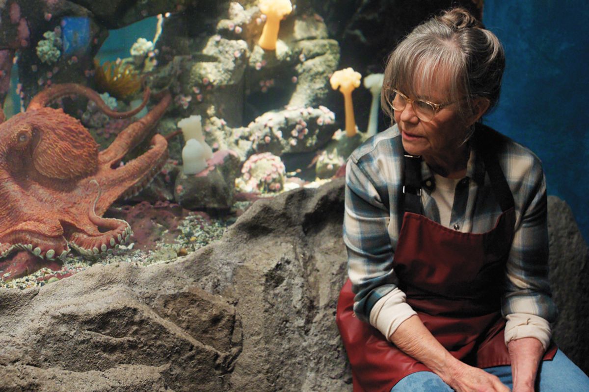 Older woman in red apron sits by a large fish tank housing a red Giant Octopus