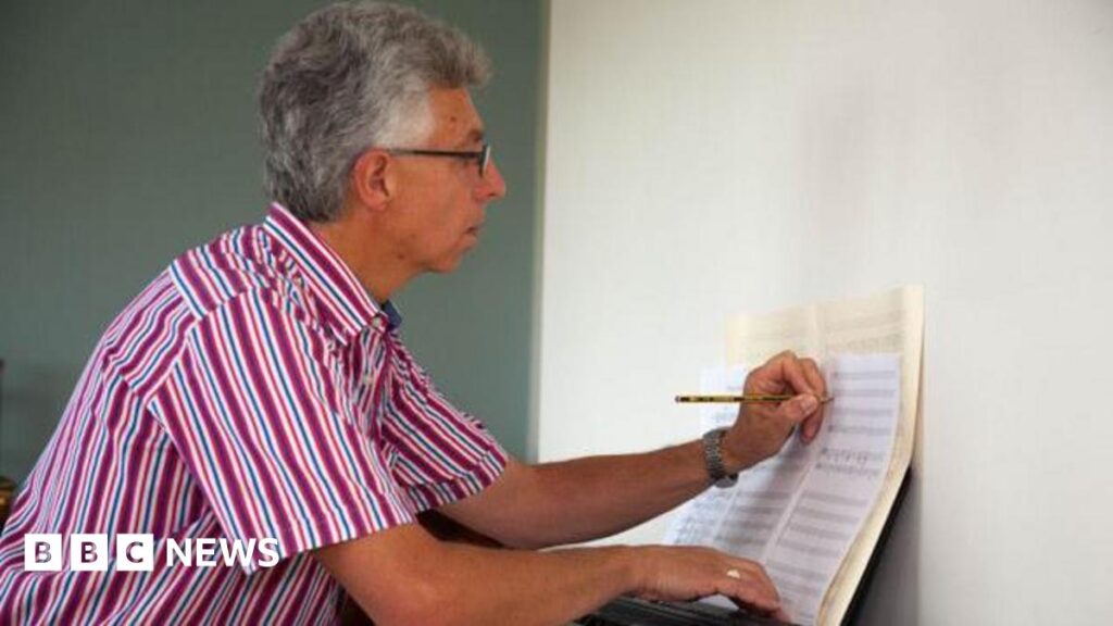 Kent composer Nick Drewe pictured composing music. Nick has graying hair and wears glasses. He is wearing a pink, blue and white striped shirt and is writing on a music sheet.