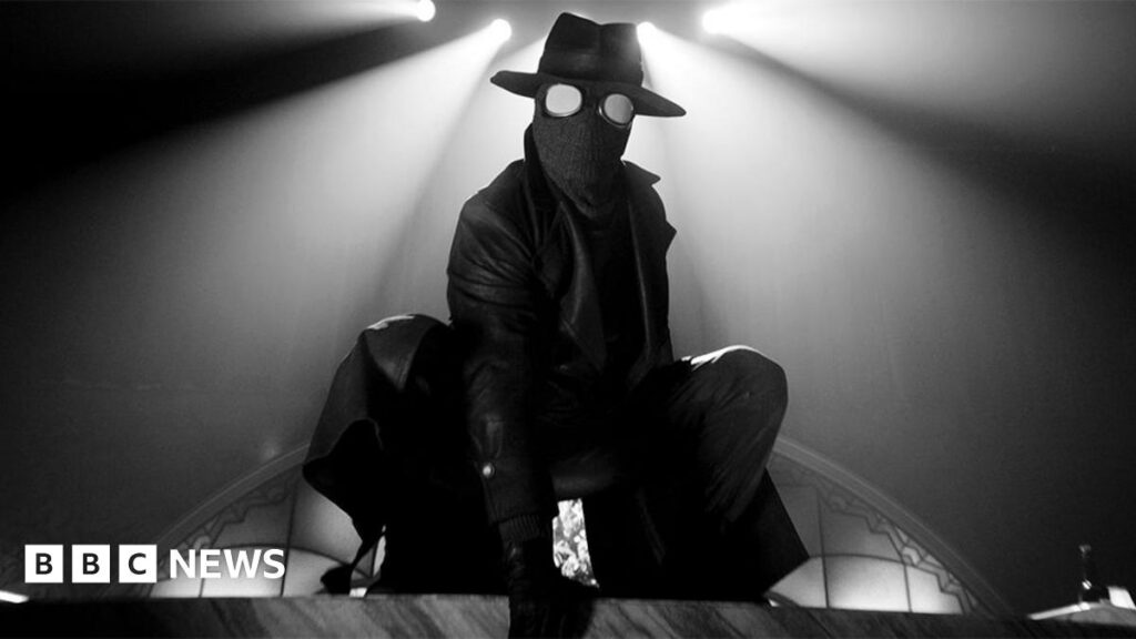 A black and white image of a character called The Spider, wearing a round hat and mask, with a leather jacket, with a white spotlight above him.