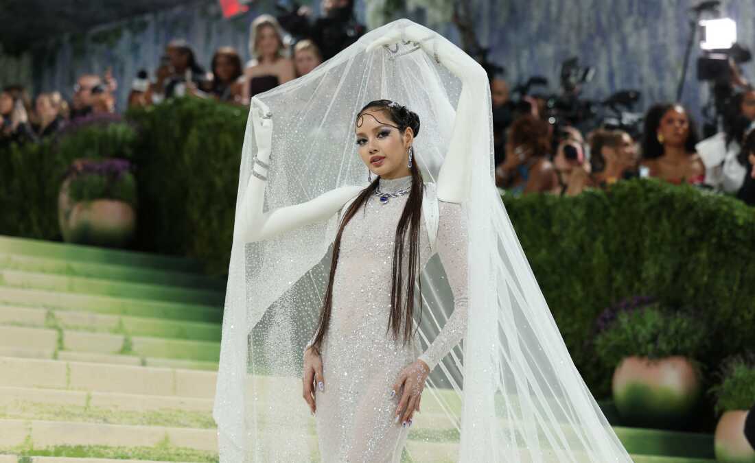 Singer Lisa arrives for the 2026 Met Gala. She is wearing a sheer, white gown with two white arms attached to her holding up a sheer, white veil. 