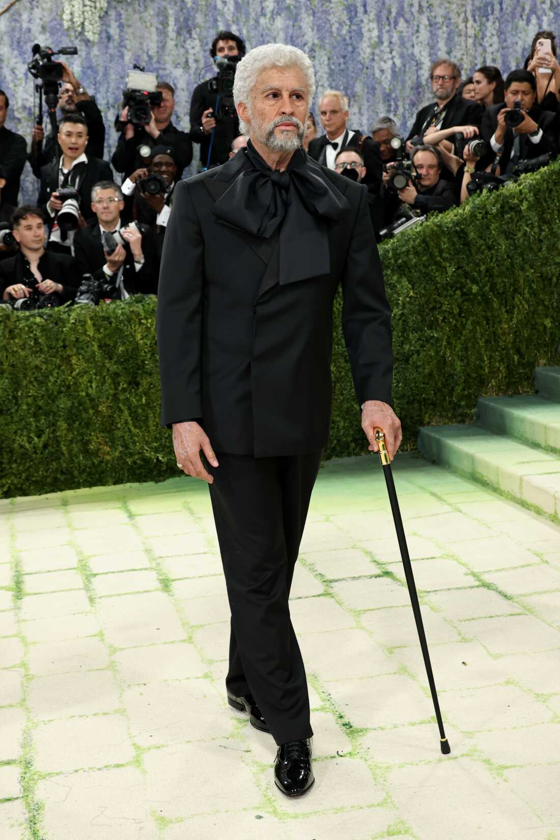 Bad Bunny attends the 2026 Met Gala. He is dressed as an old man. He is wearing a black suit and using a cane.