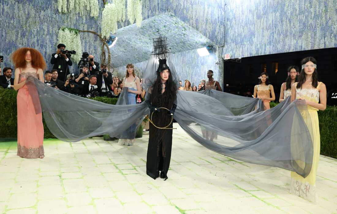 Madonna attends the 2026 Met Gala. She is wearing a black suit and a headpiece that looks like a wooden ship. There is gray fabric connected to the headpiece that is held by six models wearing pastel dresses.