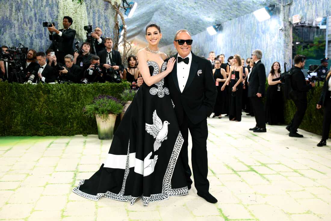 Anne Hathaway and fashion designer Michael Kors attend the 2026 Met Gala. She is wearing a black gown with a large hand reaching toward a dove. He is wearing a black tuxedo and sunglasses.