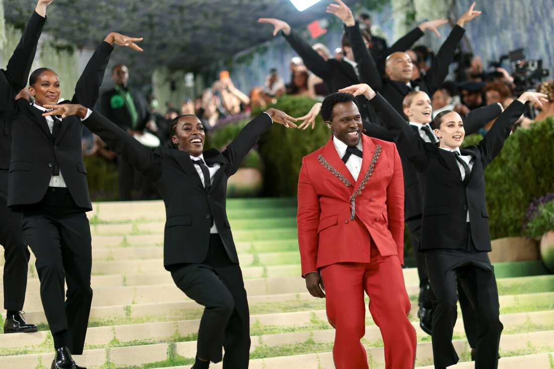 Joshua Henry performs with several backup dancers. He is wearing a red suit while the dancers are wearing black suits.