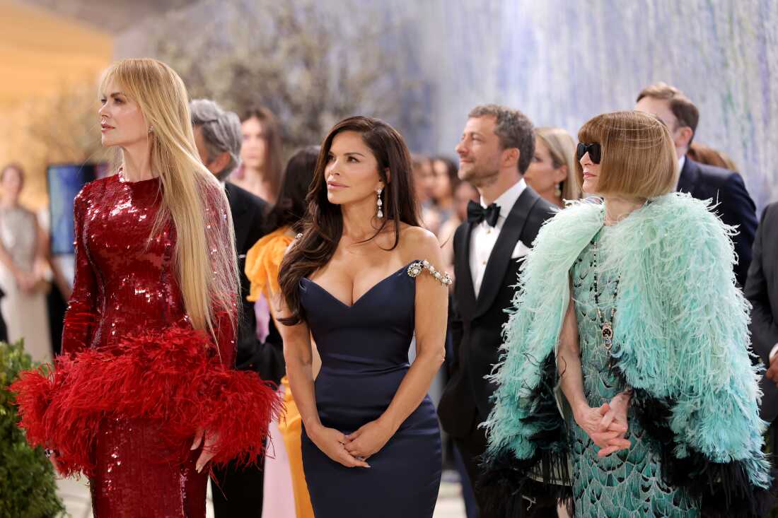 Hosts Nicole Kidman (left), Lauren Sánchez Bezos, and Anna Wintour attend the 2026 Met Gala. Kidman is wearing a red sequined gown with red feathers, Sánchez Bezos is wearing a navy gown and Wintour is wearing a light blue feathered jacket over a beaded gown.