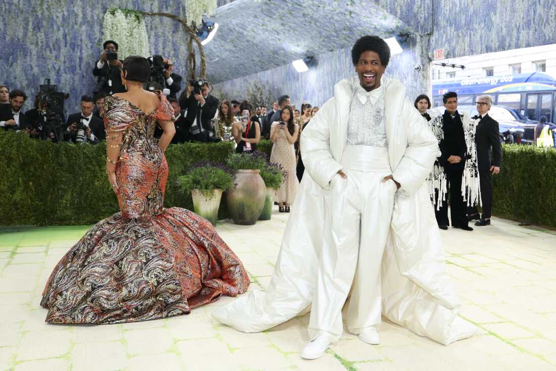 Musician Jon Batiste arrives for the 2026 Met Gala. His wife, author Suleika Jaouad is on the left. He is wearing a white suit with a white oversized, floor-length puffer jacket. She is facing away from the camera and wearing a red patterned gown.