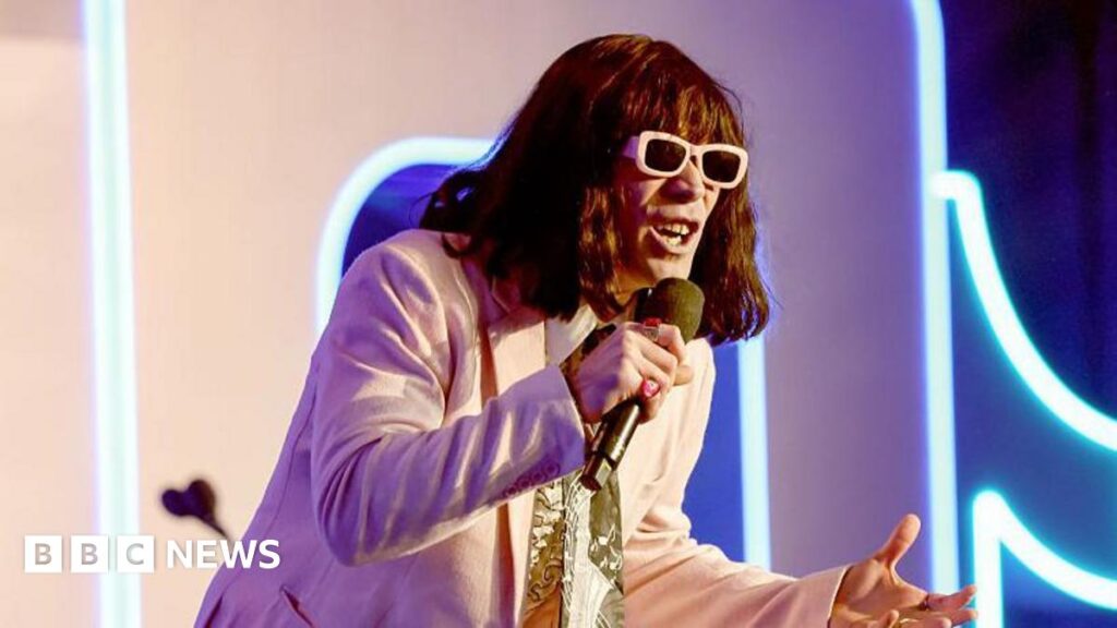 Mr Fantasy performs onstage, wearing pastel pink suit and matching sunglasses. He wears a green scarf, bright pink rings and a short brown wig with a fringe