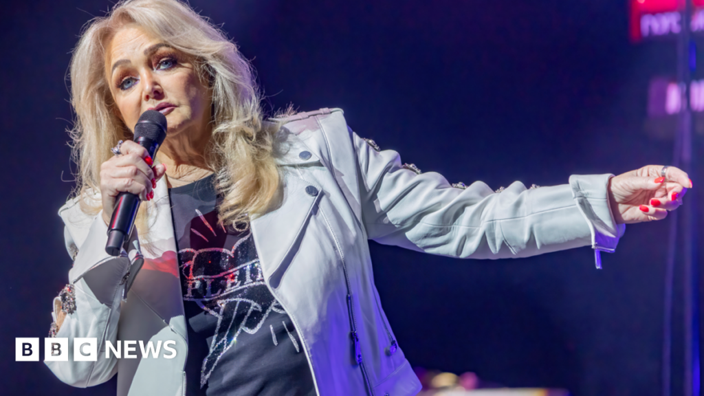 Bonnie Tyler is wearing a jacket and a black t shirt, while holding a microphone and singing.